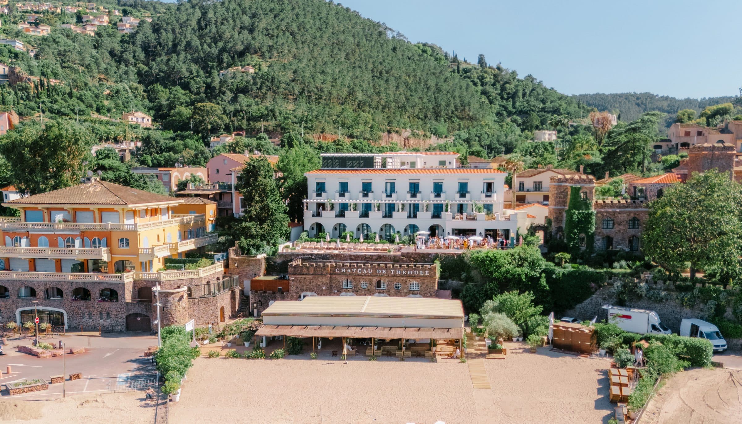 A turquoise bay and beachfront château from above, umbrellas on the sand