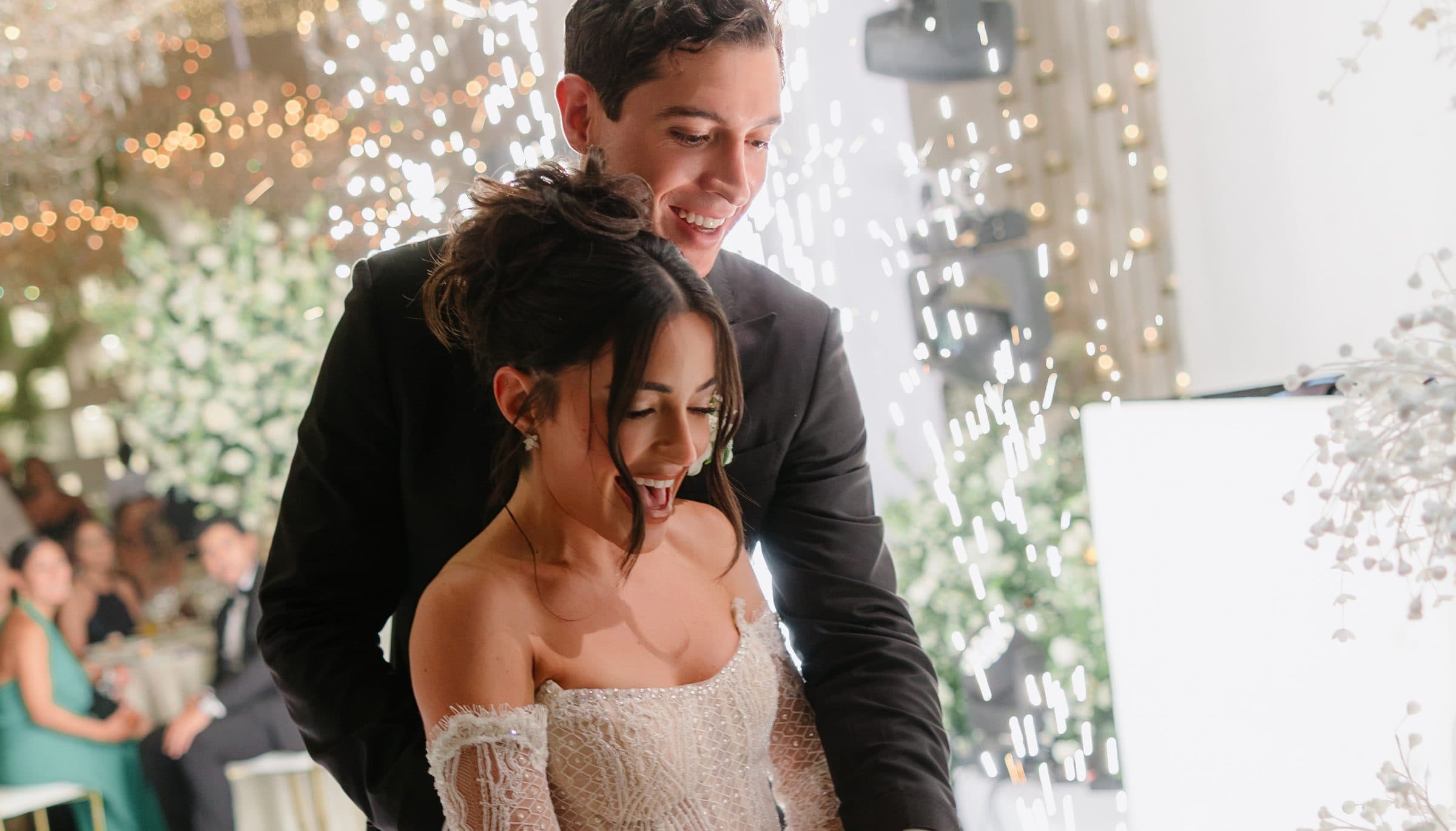 A couple cutting their wedding cake under sparkler light, ivory lace and warm skin